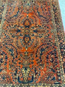 Azerbaijan Tappeti Milano Tappeto decorato in stile persiano arancione e blu con intricati motivi floreali e di viti, delimitati da una cornice geometrica. Nell'angolo in alto a destra compare il logo di una piccola foglia multicolore.