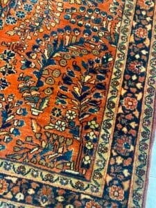 Azerbaijan Tappeti Milano Primo piano di un tappeto decorato con motivi arancioni e neri, con motivi floreali e un bordo decorativo, con il logo di una foglia d'acero colorata nell'angolo in alto a destra.