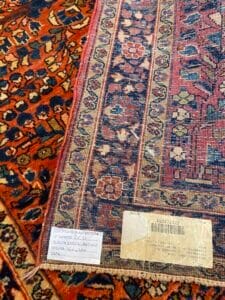 Azerbaijan Tappeti Milano Primo piano di due tappeti a motivi sovrapposti. Un tappeto ha un disegno floreale blu e arancione, mentre l'altro ha un bordo geometrico e un'etichetta di autenticità con dettagli scritti a mano e codici a barre attaccata all'angolo.