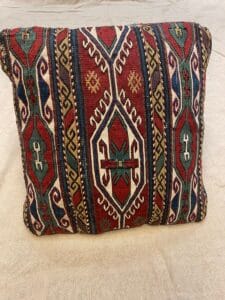 Azerbaijan Tappeti Milano Cuscino rettangolare con motivo geometrico di ispirazione tribale in rosso, verde, blu e beige. Il cuscino presenta una piccola decorazione a forma di farfalla nell'angolo in alto a destra e poggia su uno sfondo in tessuto beige.