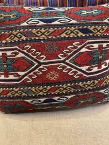 Azerbaijan Tappeti Milano Cuscino con intricati motivi geometrici in rosso, bianco, verde e blu, con disegni tradizionali di ispirazione tribale. Il bordo superiore presenta strisce multicolori e un piccolo logo di foglie e fiori nell'angolo superiore destro.