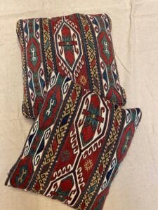 Azerbaijan Tappeti Milano Due cuscini decorativi con motivi geometrici colorati e tradizionali in rosso, verde, blu e bianco sono disposti su una superficie di tessuto beige. Un fiore multicolore e il logo "A" compaiono nell'angolo in alto a destra.