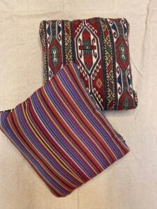 Azerbaijan Tappeti Milano Due cuscini quadrati su una superficie beige; uno presenta un motivo a righe colorate, l'altro un disegno geometrico in rosso, verde e bianco. Nell'angolo in alto a destra si trova il logo di una foglia multicolore.