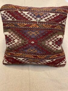 Azerbaijan Tappeti Milano Cuscino quadrato con motivo geometrico di ispirazione tribale nei toni del rosso, del bianco, del verde e del marrone, posto su una superficie in tessuto beige. Nell'angolo in alto a destra è presente un logo colorato con una foglia e la scritta "21".