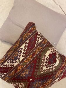 Azerbaijan Tappeti Milano Due cuscini su una superficie chiara: uno è beige semplice, l'altro presenta un motivo geometrico colorato e intricato in rosso, marrone, bianco e verde. Nell'angolo in alto a destra compare il logo di una foglia multicolore e della lettera "A".