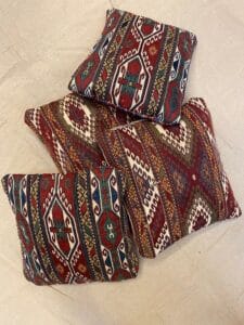 Azerbaijan Tappeti Milano Tre cuscini decorativi con motivi geometrici tradizionali nei toni del rosso, verde, crema e blu sono disposti su una superficie beige. I cuscini presentano intricati disegni di ispirazione etnica.