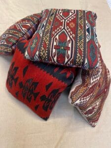 Azerbaijan Tappeti Milano Tre cuscini in tessuto colorato con motivi geometrici tradizionali in rosso, nero, verde e crema sono impilati su una superficie beige. Un cuscino presenta il logo di una farfalla multicolore nell'angolo.