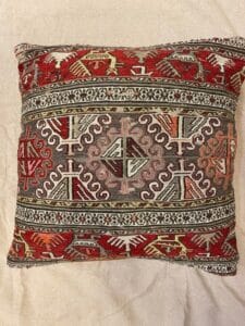 Azerbaijan Tappeti Milano Cuscino decorativo quadrato con un motivo geometrico e tribale tradizionale nei colori rosso, arancione, grigio e bianco, posto su uno sfondo in tessuto beige. Nell'angolo in alto a destra compare il logo di una foglia colorata e di un cuore.