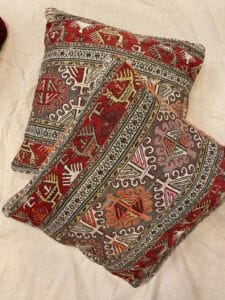 Azerbaijan Tappeti Milano Due cuscini quadrati con intricati e colorati motivi geometrici e tribali nei toni del rosso, bianco, rosa e giallo sono posizionati su una superficie chiara. Un piccolo logo multicolore compare nell'angolo in alto a destra.