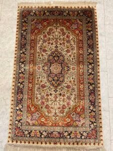 Azerbaijan Tappeti Milano Tappeto persiano rettangolare con intricati motivi floreali in rosso, crema e blu navy, delimitati da disegni dettagliati. Il tappeto è posizionato su un pavimento piastrellato chiaro, con frange sui bordi più corti.