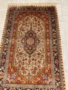 Azerbaijan Tappeti Milano Un tappeto persiano rettangolare con intricati motivi floreali e geometrici nei toni del rosso, del crema, del nero e dell'oro giace su un pavimento piastrellato chiaro. Nell'angolo in alto a destra compare il logo di una foglia colorata.