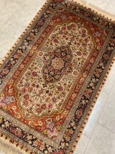 Azerbaijan Tappeti Milano Un tappeto rettangolare in stile persiano con intricati motivi floreali nei toni del rosso, del crema e del nero giace su un pavimento piastrellato. Il tappeto presenta bordi ornati e smerlati a ogni estremità.