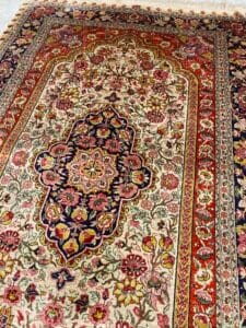 Azerbaijan Tappeti Milano Tappeto persiano dettagliato e decorato con intricati motivi floreali nei toni del rosso, del blu, del crema e dell'oro, delimitati da disegni geometrici. Nell'angolo in alto a destra è visibile il logo di una foglia colorata e moderna.