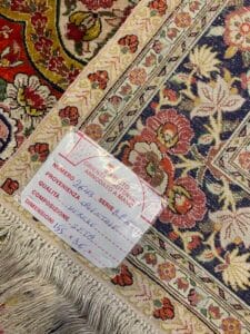 Azerbaijan Tappeti Milano Primo piano di un tappeto a fantasia con un'etichetta che riporta dettagli scritti a mano in italiano, tra cui dimensioni e qualità, vicino al bordo sfrangiato. Nell'angolo in alto a destra compare il logo di una foglia colorata.