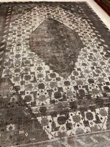 Azerbaijan Tappeti Milano Un tappeto con motivo bianco e nero, un grande motivo centrale a forma di diamante e un logo a forma di foglia colorata nell'angolo.