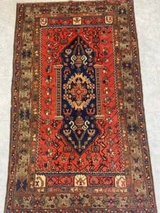 Azerbaijan Tappeti Milano Tappeto orientale rettangolare con medaglione centrale, con motivi rossi, blu navy e verde oliva, motivi geometrici e un bordo decorativo. Nell'angolo superiore destro compare il logo di una foglia colorata.