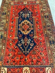Azerbaijan Tappeti Milano Tappeto tradizionale in stile persiano con audaci motivi geometrici e floreali nei toni del rosso, del blu e del crema, con intricati disegni sui bordi e un medaglione centrale. Un logo multicolore compare nell'angolo superiore destro.