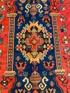 Azerbaijan Tappeti Milano Un tappeto colorato e intricato con disegni geometrici e floreali rossi, blu e arancioni, con motivi simmetrici e un medaglione centrale. Nell'angolo in alto a destra compare una piccola farfalla multicolore.