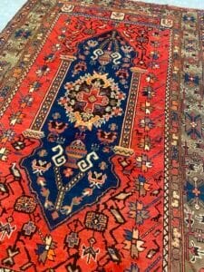 Azerbaijan Tappeti Milano Tappeto persiano colorato e decorato con un medaglione centrale blu scuro con intricati motivi floreali e geometrici, circondato da toni rossi e terrosi con bordi decorativi dettagliati. Un piccolo logo colorato si trova nell'angolo superiore destro.