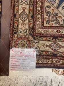 Azerbaijan Tappeti Milano Primo piano di un tappeto a motivi geometrici e floreali. Sul tappeto è apposta un'etichetta in italiano che riporta dettagli quali origine, qualità, composizione, dimensioni e prezzo. Sul bordo del tappeto sono visibili delle frange.