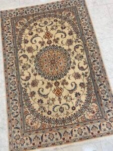 Azerbaijan Tappeti Milano Tappeto rettangolare decorato in stile persiano con intricati motivi floreali e geometrici nei toni del beige, blu, marrone e nero, esposto su un pavimento piastrellato. Un logo colorato appare nell'angolo in alto a destra.