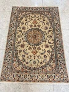 Azerbaijan Tappeti Milano Tappeto rettangolare in stile persiano con un intricato motivo floreale e di viti nei toni del beige, del blu e del marrone, caratterizzato da un medaglione centrale e da bordi decorativi, posto su un pavimento piastrellato chiaro.