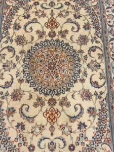 Azerbaijan Tappeti Milano Un tappeto beige e marrone chiaro presenta un intricato motivo floreale simmetrico con dettagli blu e color ruggine. Il bordo presenta motivi coordinati. Nell'angolo in alto a destra si trova un logo astratto e colorato.