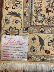 Azerbaijan Tappeti Milano Primo piano di un tappeto a fantasia con un cartellino che riporta dettagli scritti a mano in italiano su origine, qualità, composizione, dimensioni e prezzo. Il tappeto presenta disegni floreali blu, beige e marroni.