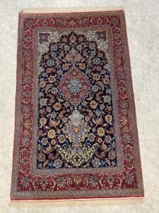 Azerbaijan Tappeti Milano Tappeto persiano rettangolare con un intricato motivo floreale nei toni del blu, del rosso, dell'avorio e dell'oro, caratterizzato da un medaglione centrale dettagliato, delimitato da una spessa fascia rossa, esposto su un pavimento piastrellato chiaro.