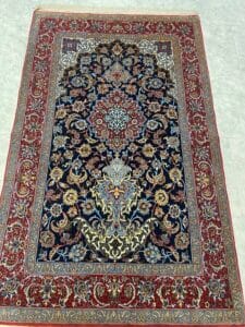 Azerbaijan Tappeti Milano Tappeto decorativo in stile persiano con intricati motivi floreali in rosso, blu, oro e crema, bordato con disegni ornamentali, esposto su un pavimento piastrellato chiaro. Un logo colorato si trova nell'angolo in alto a destra.