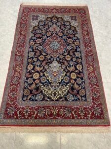 Azerbaijan Tappeti Milano Un tappeto persiano rettangolare con un intricato motivo floreale nei toni del rosso, del blu, del crema e dell'oro, caratterizzato da un medaglione centrale e da bordi ornati, steso su un pavimento di piastrelle chiare.