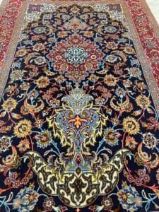 Azerbaijan Tappeti Milano Un tappeto persiano ornato con intricati motivi floreali e geometrici in rosso, blu, oro e crema su sfondo scuro. Nell'angolo in alto a destra compare il logo di una foglia colorata.