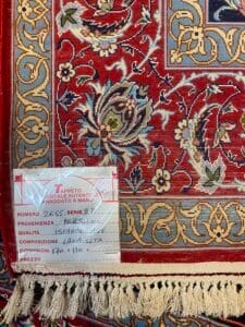Azerbaijan Tappeti Milano Primo piano di un dettagliato tappeto persiano con motivi floreali in rosso, blu e beige. Un'etichetta allegata fornisce specifiche in italiano, tra cui origine, qualità e dimensioni. Il tappeto presenta bordi sfrangiati.