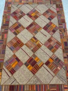 Azerbaijan Tappeti Milano Un tappeto con motivo patchwork caratterizzato da quadrati e rettangoli nei toni della terra e colori vivaci, con un simbolo decorativo nell'angolo in alto a destra.