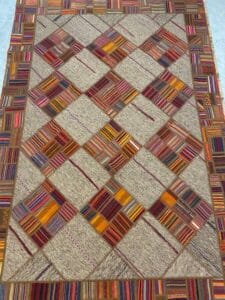 Azerbaijan Tappeti Milano Un tappeto patchwork con motivi geometrici e quadrati multicolori. Nell'angolo in alto a destra è visibile un logo colorato con un motivo a foglia e vortice.