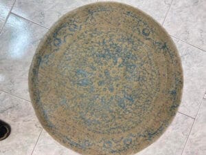 Azerbaijan Tappeti Milano Tappeto beige rotondo con motivi blu su pavimento piastrellato. Disegno di foglie colorate e scritta nera nell'angolo in alto a destra.