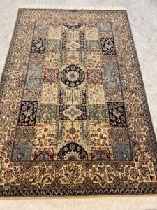 Azerbaijan Tappeti Milano Su un pavimento di piastrelle chiare è esposto un tappeto rettangolare in stile persiano con motivi floreali e geometrici nei toni del beige, marrone, rosso, blu e verde. Nell'angolo in alto a destra è presente un logo colorato.