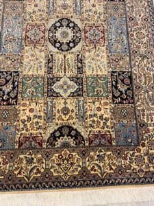 Azerbaijan Tappeti Milano Un tappeto dettagliato in stile persiano con intricati motivi floreali e geometrici nei toni del blu, del crema e del rosso. Il logo di una farfalla colorata con ali rosse, verdi, arancioni e gialle si trova nell'angolo superiore destro. La frangia è visibile nella parte inferiore.