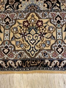 Azerbaijan Tappeti Milano Primo piano di un tappeto ornato con intricati motivi floreali in beige, marrone e blu, con frange bianche in basso e un logo a forma di farfalla colorata nell'angolo in alto a destra.