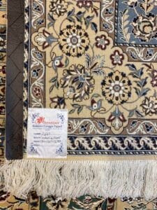 Azerbaijan Tappeti Milano Primo piano di un tappeto azerbaigiano con motivi floreali intricati nei toni del blu, del beige e del marrone, con frange bianche sul fondo e un'etichetta con i dettagli del tappeto in un angolo.