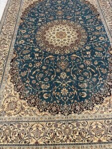 Azerbaijan Tappeti Milano Tappeto decorativo in stile persiano con motivi floreali ornamentali nei toni del crema e del blu scuro, caratterizzato da un dettagliato medaglione circolare centrale e da bordi intricati. Nell'angolo superiore destro compare un logo astratto e colorato.