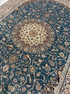 Azerbaijan Tappeti Milano Tappeto ornato in stile persiano con un intricato motivo floreale in beige e oro su uno sfondo blu intenso. Nell'angolo superiore destro del tappeto è visibile il logo di una stella colorata a più punte.