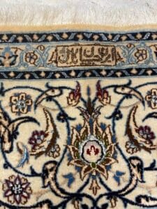 Azerbaijan Tappeti Milano Primo piano di un tappeto con disegni floreali nei colori blu, marrone e crema. C'è una sezione rettangolare con scritte in arabo e un logo floreale colorato nell'angolo in alto a destra.