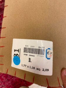 Azerbaijan Tappeti Milano Primo piano di un'etichetta di tessuto con numeri, un codice a barre e un adesivo blu su un tessuto beige e rosso. Un logo a forma di foglia colorata è nell'angolo in alto a destra.