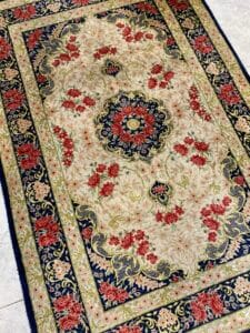 Azerbaijan Tappeti Milano Un tappeto rettangolare in stile persiano con intricati motivi floreali e un medaglione centrale nei colori rosso, crema e blu navy. Un logo colorato a forma di farfalla è su un angolo.
