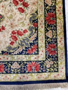 Azerbaijan Tappeti Milano Primo piano di un tappeto con motivo floreale, bordo blu navy e oro, caratterizzato da fiori rossi e un motivo decorativo a farfalla in un angolo.