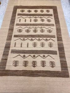 Azerbaijan Tappeti Milano Un tappeto beige e marrone con motivi geometrici a zigzag, rombi e linee. Un piccolo logo multicolore si trova nell'angolo in alto a destra.