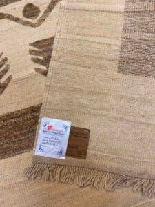 Azerbaijan Tappeti Milano Primo piano di un tappeto beige intrecciato con bordo sfrangiato. Sono visibili due etichette, una floreale e una con testo. Un logo colorato è nell'angolo in alto a destra.