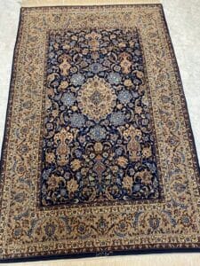 Azerbaijan Tappeti Milano Tappeto rettangolare in stile persiano con intricati motivi floreali e di viti nei toni del blu, del beige e dell'oro e un grande medaglione centrale. Nell'angolo in alto a destra dell'immagine compare il logo di una foglia colorata.