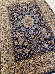 Azerbaijan Tappeti Milano Un grande tappeto persiano ornato con intricati motivi floreali nei toni del beige, dell'oro e del blu su uno sfondo blu scuro. Un logo colorato con foglie sovrapposte è visibile nell'angolo in alto a destra.